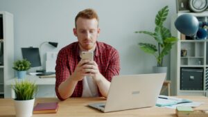 Small business owner looking concerned while checking bank balance on a phone at a desk